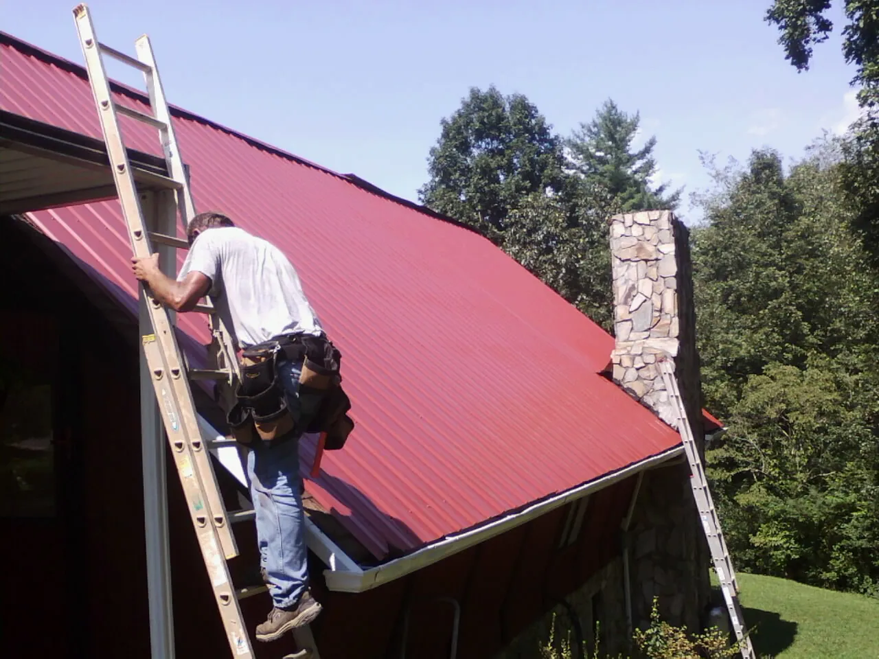 Roofer on a ladder beside a red metal roof and stone chimney — Soffit & Fascia Repair by Roofing Brothers in Medford